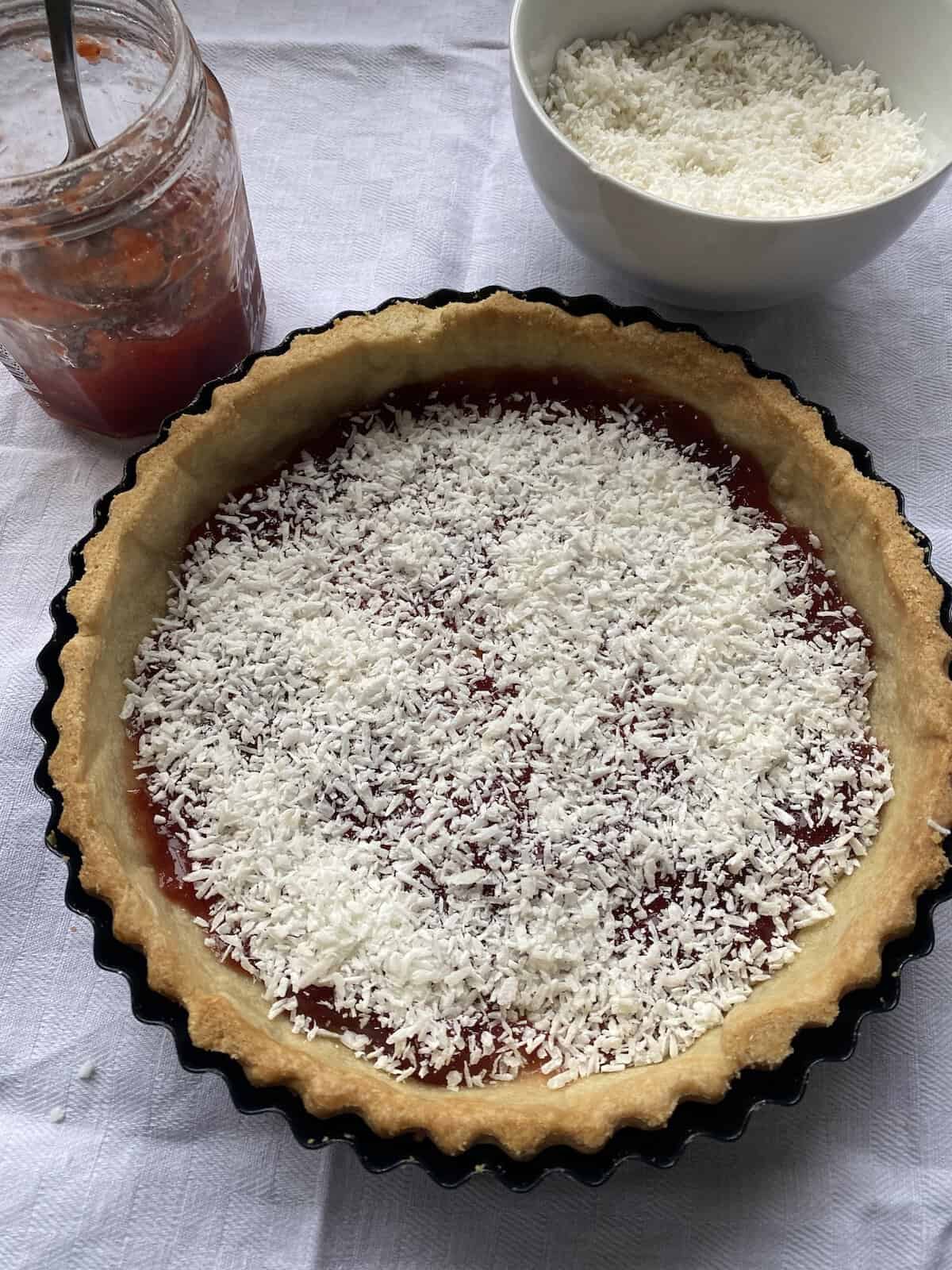 Jam and coconut spread over the base of a baked pastry tart.
