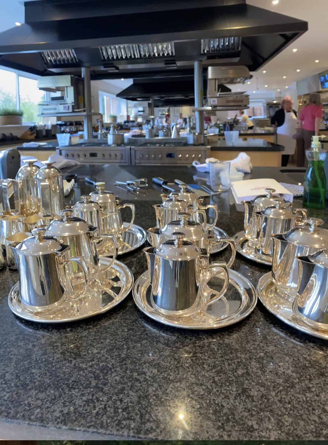 Bettys Cookery School kitchen, with rows of silver tea pots in the foreground. Collection of Silver Tea pots on a counter top.