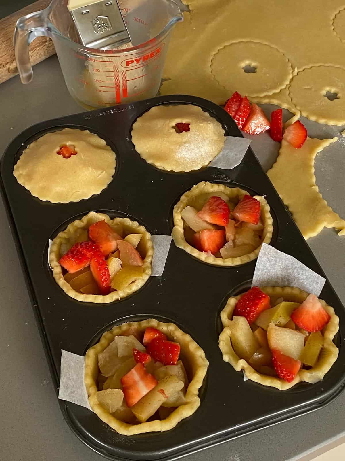 six unbaked fruit pies in a baking tray. Tray of unbaked small strawberry and apple tarts.